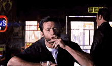 a man is smoking a cigar and drinking a glass of whiskey while sitting at a table in a bar .