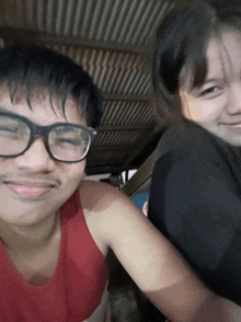a man wearing glasses and a red tank top smiles with a woman behind him