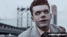 a man in a suit and tie with a joker mask on his face is standing in front of a bridge .