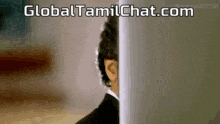 a man in a suit peeking out from behind a wall with the website global tamilchat.com written above him .