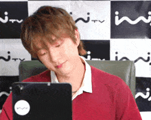 a man in a red sweater is sitting in front of a black and white checkered wall looking at a tablet .