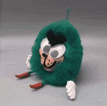 a green stuffed animal with googly eyes and red feet is sitting on a table