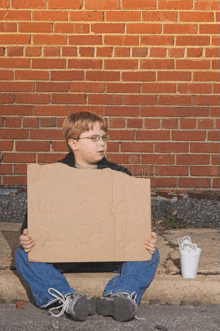 a young boy is sitting on the sidewalk holding a cardboard sign in front of a brick wall
