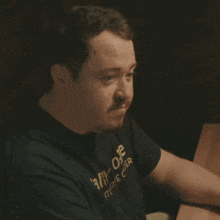 a man wearing a black shirt that says ally one is sitting at a table