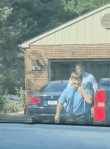 a man in a blue shirt and tie is giving a thumbs up while standing in front of a brick house .