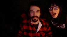 a man in a red plaid shirt and a woman in glasses are looking at the camera