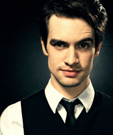a man wearing a white shirt and a black vest and tie looks at the camera