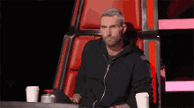 a man in a black jacket is sitting in a chair with two cups of coffee .