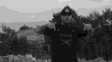 a black and white photo of a man wearing a hat and a t-shirt standing in front of a building .