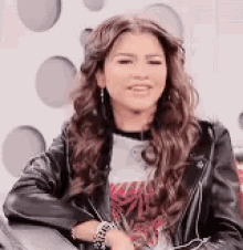 a woman wearing a leather jacket and a t-shirt is sitting on a couch and smiling .
