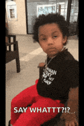 a young boy wearing a black shirt and red pants is sitting on a chair and says say whattt ?