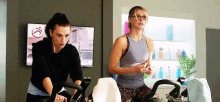 two women are riding exercise bikes in a gym with a sign on the wall that says " i "