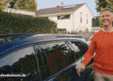 a man in an orange sweater is walking towards a blue car with a house in the background