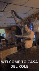 a man without a shirt is sitting at a table in a bar with the words welcome to casa del kolb above him