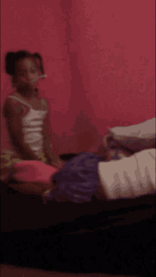 a girl in a white tank top sits on a bed with a pink wall behind her