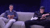 two men are sitting on a couch looking at their tablets