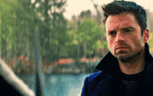 a man in a blue jacket is standing in front of a body of water with trees in the background .