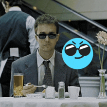 a man in a suit and tie sits at a table with a blue smiley face in the background