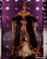 a drag queen is standing in front of a purple curtain wearing a fur coat .