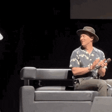 a man wearing a hat is sitting in a chair holding a microphone and clapping .