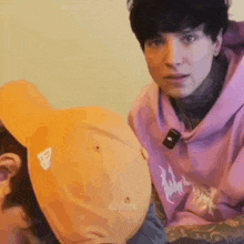 a man in a pink hoodie is sitting next to another man wearing a yellow hat .