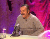 a man with a mustache is sitting at a table with a microphone and a glass of water .