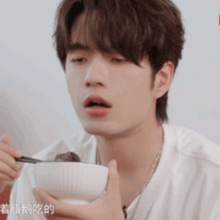 a young man in a white shirt is eating from a white bowl with chopsticks