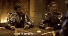 two men in armor are sitting at a table with a candle and the words c ' est le progress written on the bottom .