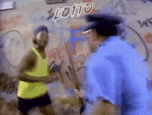 a man in a yellow tank top is standing next to a police officer in front of a wall with graffiti that says lotto