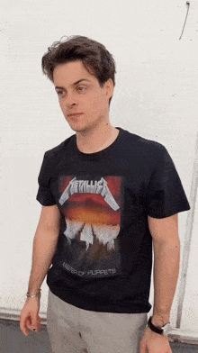 a man wearing a black metallica t-shirt stands in front of a white wall