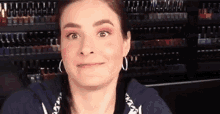 a woman wearing hoop earrings and pink eyeshadow is making a funny face in front of a shelf of nail polish .