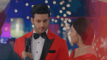 a man in a red tuxedo is talking into a microphone while a woman in a red dress looks on