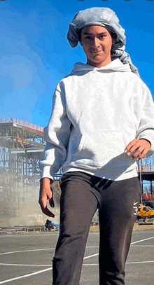 a man wearing a white hoodie and black nike sweatpants stands in front of a construction site