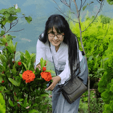 a woman wearing glasses and a black purse stands in front of a bush with red flowers