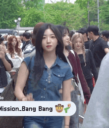 a woman in a denim shirt stands in a crowd with a mission bang sign behind her