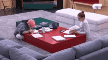 two women are sitting at a red table in a living room with the letter t on the wall