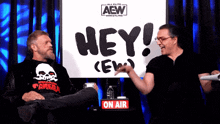 two men are sitting in front of a sign that says hey crew