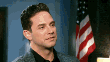 a man in a suit and black shirt stands in front of an american flag