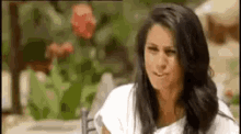 a woman with long dark hair is sitting at a table with flowers in the background .