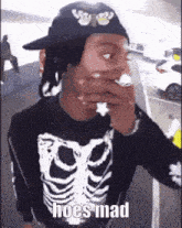 a man wearing a skeleton shirt and a hat is smoking a cigarette and says hoes mad .