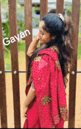 a woman in a red saree is standing in front of a wooden fence with a ponytail .