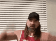 a man with long hair and a beard wearing a baseball cap and a red tank top is standing in front of a window .