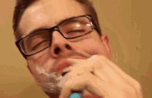 a man wearing glasses is shaving his face with a razor and foam .