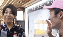 a man wearing a pink hat talks to another man in front of a cheese shop