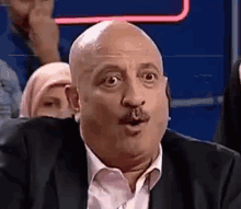 a bald man with a mustache is making a funny face while sitting in front of a group of people .
