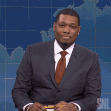 a man in a suit and tie sitting in front of a snl logo