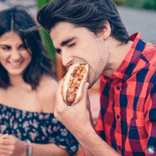 a man in a plaid shirt is eating a hot dog while a woman watches .