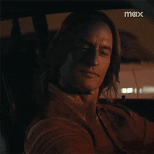 a man with long hair is sitting in the driver 's seat of a car with the word max on the bottom right
