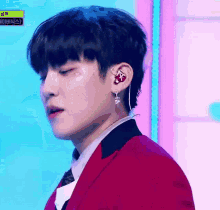 a young man wearing a red jacket and earrings is standing on a stage with his eyes closed .