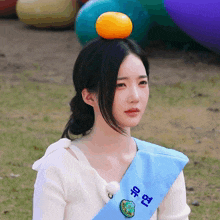 a girl wearing a sash that says ' seoul ' on it has an orange on her head